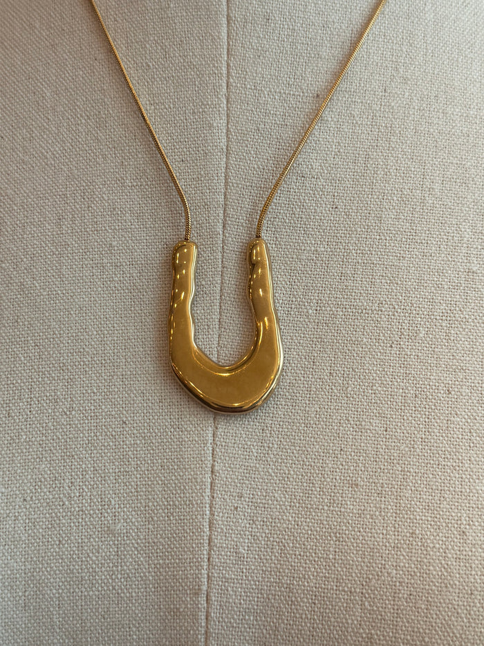 Gold wavy U pendant necklace detail with thin chain on fabric background