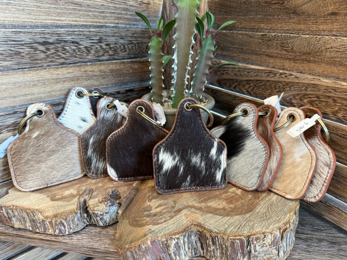 A set of western-style leather keychains or tags with cowhide-patterned backs, arranged on a rustic wood slice in front of a wooden wall.