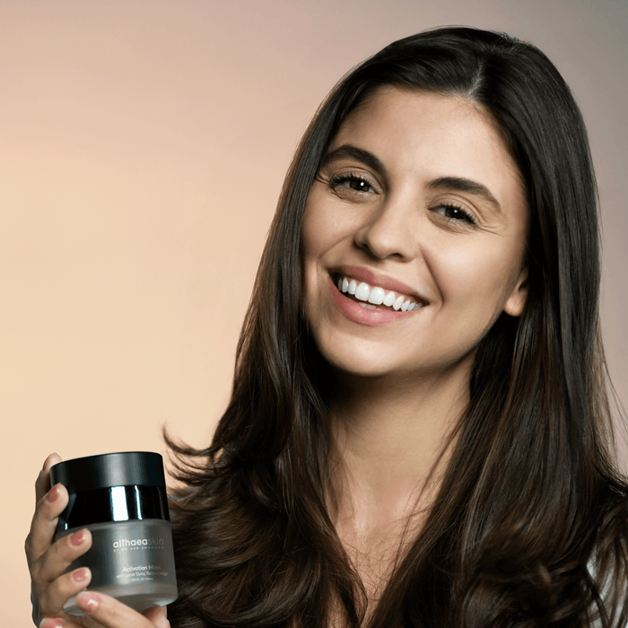 Smiling woman with long dark hair holding a jar of Activation Mask | Althaea Skin, showcasing a skincare product for enhancing skin wellness and radiance.