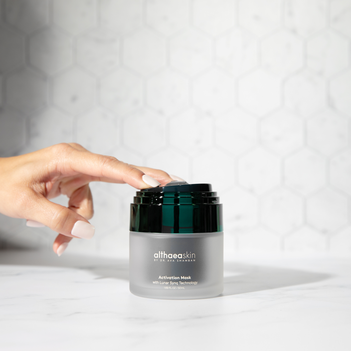 Hand with manicured nails opening a jar of Activation Mask | Althaea Skin placed on a white marble surface with a soft hexagonal pattern background, showcasing elegant skincare packaging.