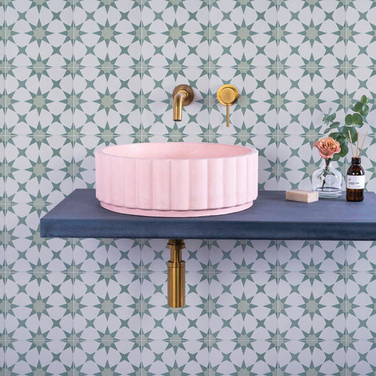 Pink sink on a blue shelf with gold fixtures against green patterned wall tiles