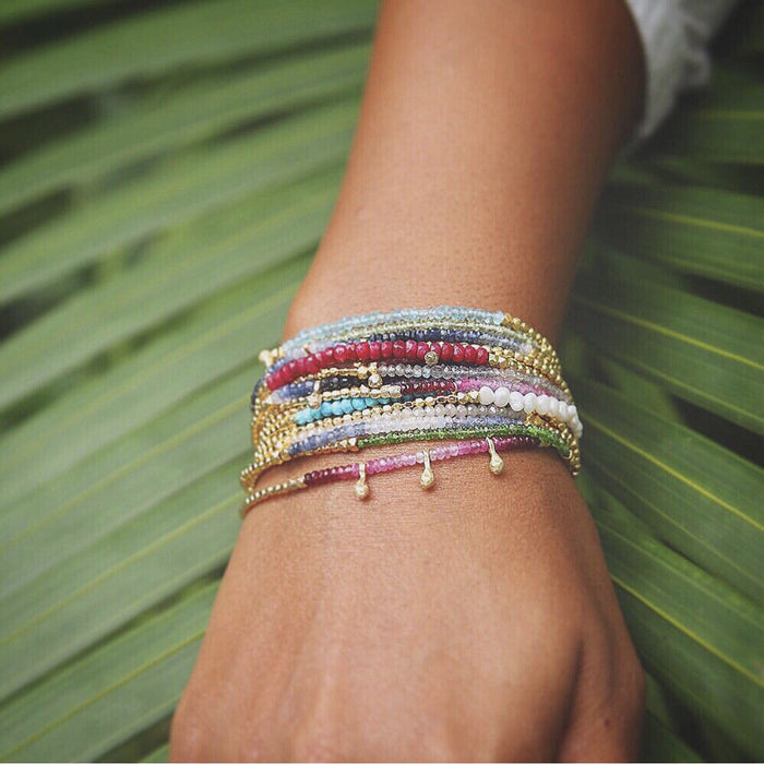 Close-up of a wrist wearing the Gemstone Fade Bracelet | Catherine Weitzman Jewelry, featuring multiple delicate strands with colorful gemstones and gold beads against a green leaf background.