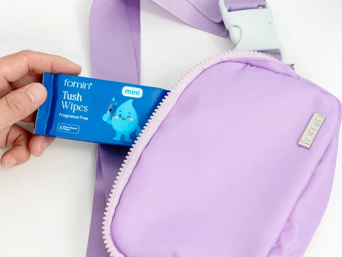 Hand holding fomin Tush Wipes next to a purple bag on a white background