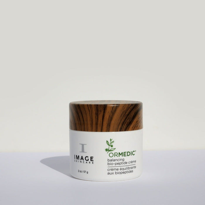 Jar of ORMEDIC balancing biopeptide crème | IMAGE Skincare with a white container and wood-textured lid, placed on a light surface with a neutral gray background.