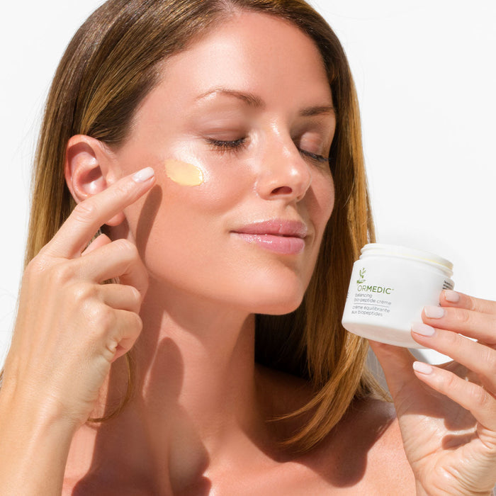 Woman with clear skin gently applies ORMEDIC balancing biopeptide crème | IMAGE Skincare to her cheek, holding the white jar in her hand against a bright background.