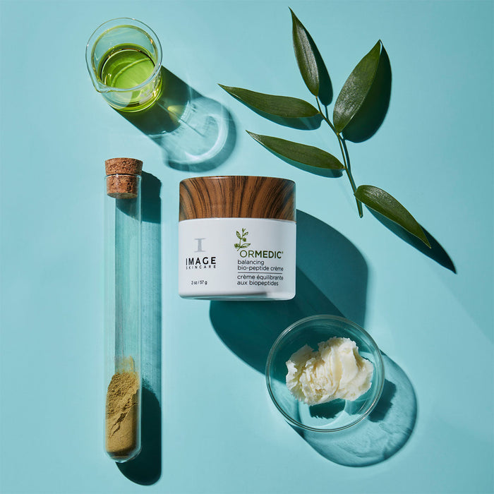 ORMEDIC balancing biopeptide crème | IMAGE Skincare jar with natural ingredients including green oil, powder in a glass tube, plant leaves, and a bowl of cream on a blue background, highlighting skincare and wellness.