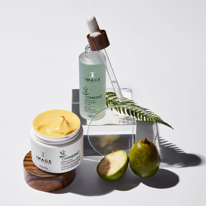 Open jar of yellow ORMEDIC balancing biopeptide crème | IMAGE Skincare on wooden lid next to clear dropper bottle with green serum, fresh green fruit halves, and a fern leaf on a clear stand, skincare product arrangement.