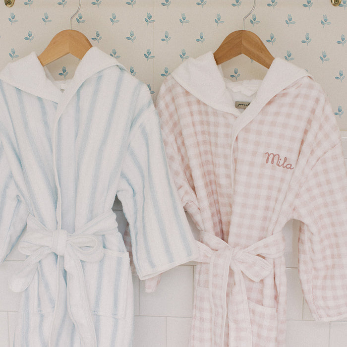 Two hooded bathrobes hanging on wooden hangers against patterned wallpaper. The left robe is white with light blue vertical stripes and a white hood.