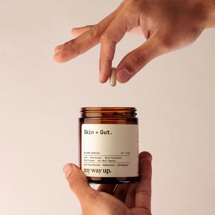 Hand holding a jar labeled 'Skin + Gut' with a pill above it on a beige background